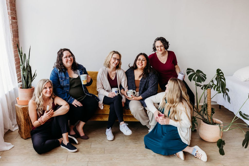 All of the members of Welcome Baby KC sitting on a couch together with coffee mugs in their hands.