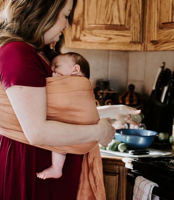 A newborn baby being worn in a wrap by a postpartum doula while she cooks a meal